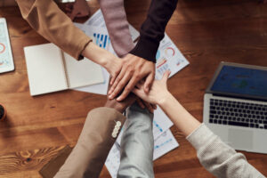 Fünf Personen legen ihre Hände in einer Geste der Teamarbeit über einem Holztisch zusammen, der mit Dokumenten und Diagrammen übersät ist, als würden sie Rätseltouren planen oder ihr nächstes Escape-Room-Abenteuer brainstormen.