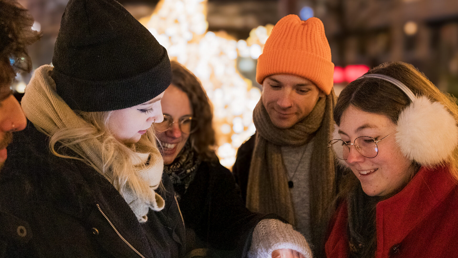 Eine Gruppe von fünf Personen in Winterkleidung lächelt und schaut gemeinsam auf ein Tablet im nächtlichen Freien, umgeben von festlichen Lichtern - sie genießen spannende Rätseltouren als Teil ihres Outdoor-Touren-Abenteuers inmitten von Weihnachtsdekoration.