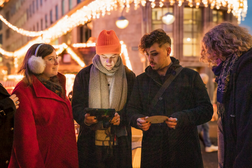 Vier Personen in Winterkleidung stehen zusammen auf einem mit Lichterketten geschmückten Markt im Freien. Sie scheinen sich zu unterhalten, vielleicht planen sie eine der örtlichen Rätseltouren - ein Outdoor-Escape-Room-Abenteuer mit einem Tablet und Hinweisen.