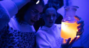 Zwei Personen untersuchen eine leuchtende gelbe Flüssigkeit in einem Glasgefäß bei blauem Licht. Eine Person hält eine Taschenlampe, um das Glas besser sehen zu können, während beide neugierig und erstaunt wirken.