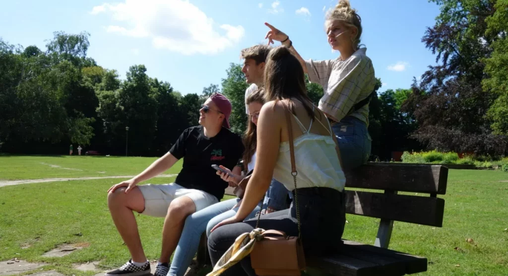 Vier junge Leute sitzen bei sonnigem Wetter auf einer Parkbank, mit Bäumen und Gras im Hintergrund. Eine Person zeigt in die Ferne, während die anderen in diese Richtung blicken. Eine braune Handtasche liegt neben ihnen.