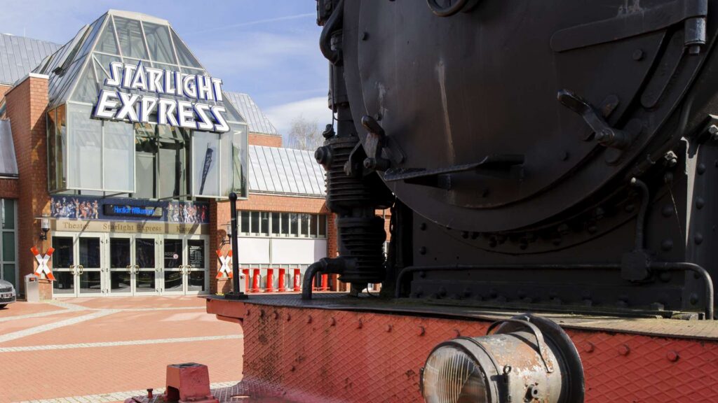 Eine große, schwarze Dampflokomotive steht vor dem Eingang des Starlight Express Theaters, das eine Glasfassade und ein auffälliges Starlight Express-Schild über den Türen hat.